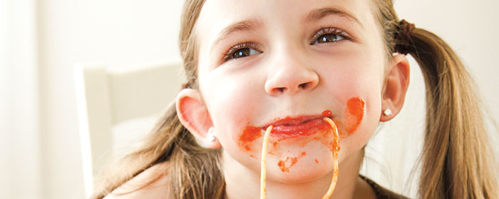A smiling girl with piece of spaghetti hanging from her mouth and sauce on her cheeks