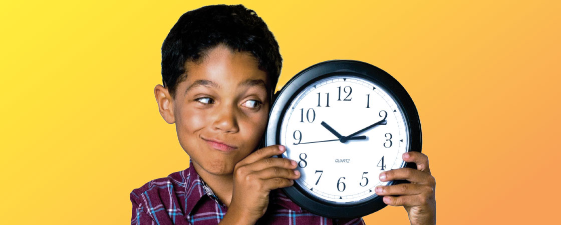 A boy wearing a plaid shirt looking at the round clock he is holding up next to his face