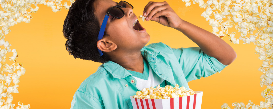 Kid eating popcorn and wearing 3-D glasses against a yellow popcorn background