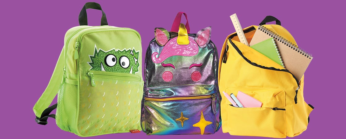 Three children's backpacks: a green monster design, a holographic unicorn, and a yellow bag filled with school supplies